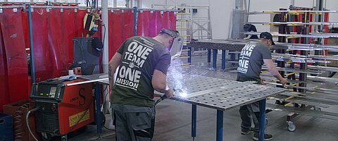 Arbeiter schweißt Metall, zweiter Arbeiter verräumt Metall